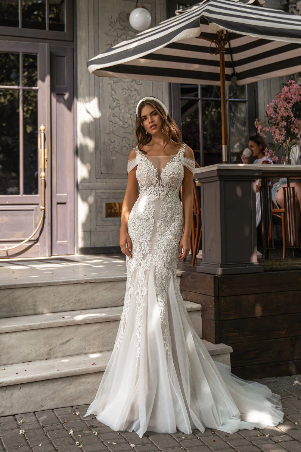 Woman in a white lace wedding dress standing outdoors near a building entrance.