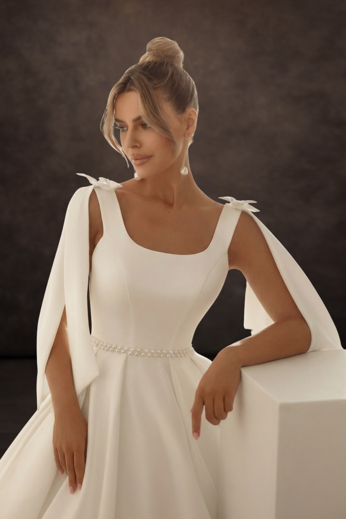 Woman in a off white wedding dress with bows on her shoulders sitting on a white surface against a dark background