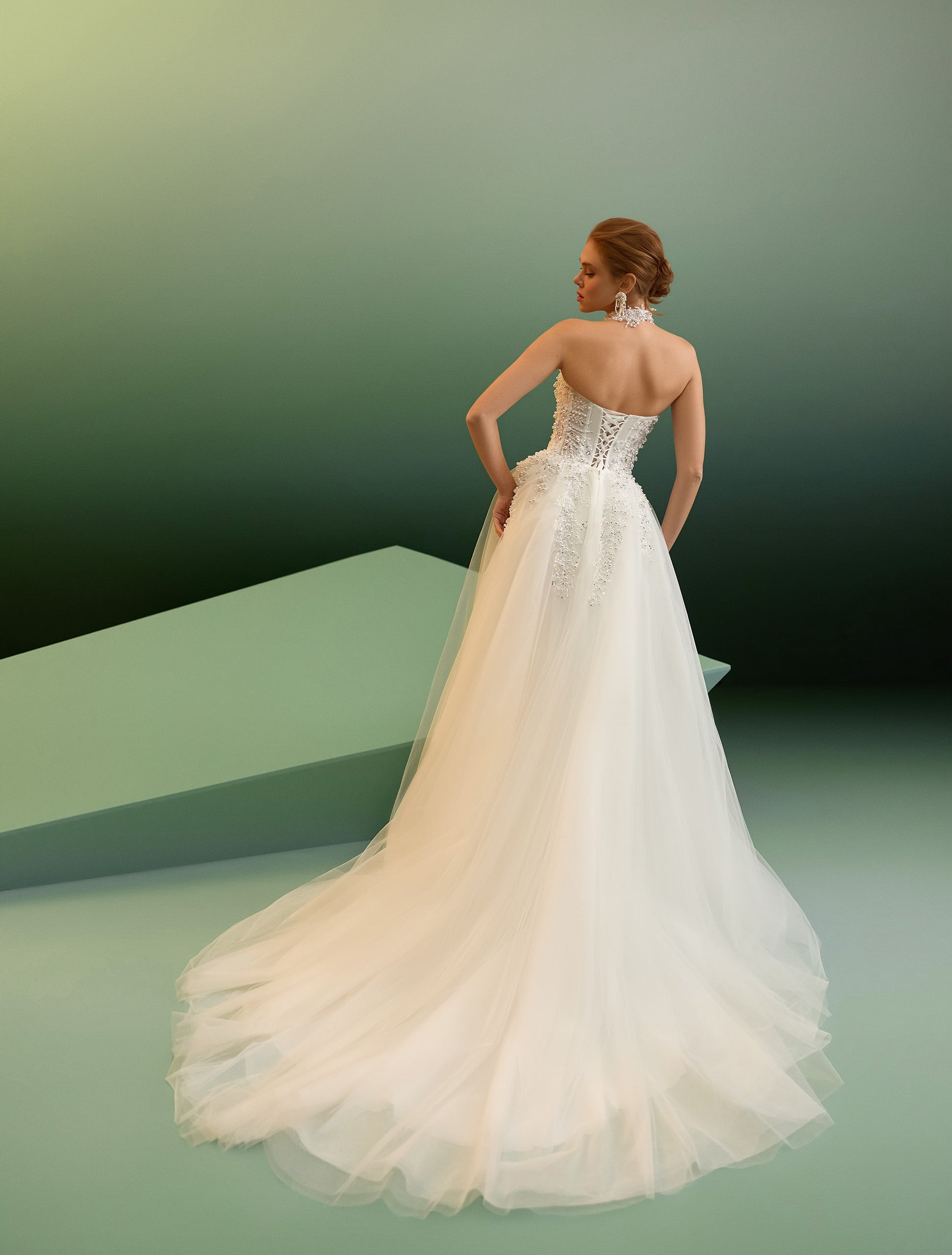 A woman wearing a strapless white wedding gown with a fitted lace bodice and a full tulle train, photographed from the back against a soft green gradient studio background.