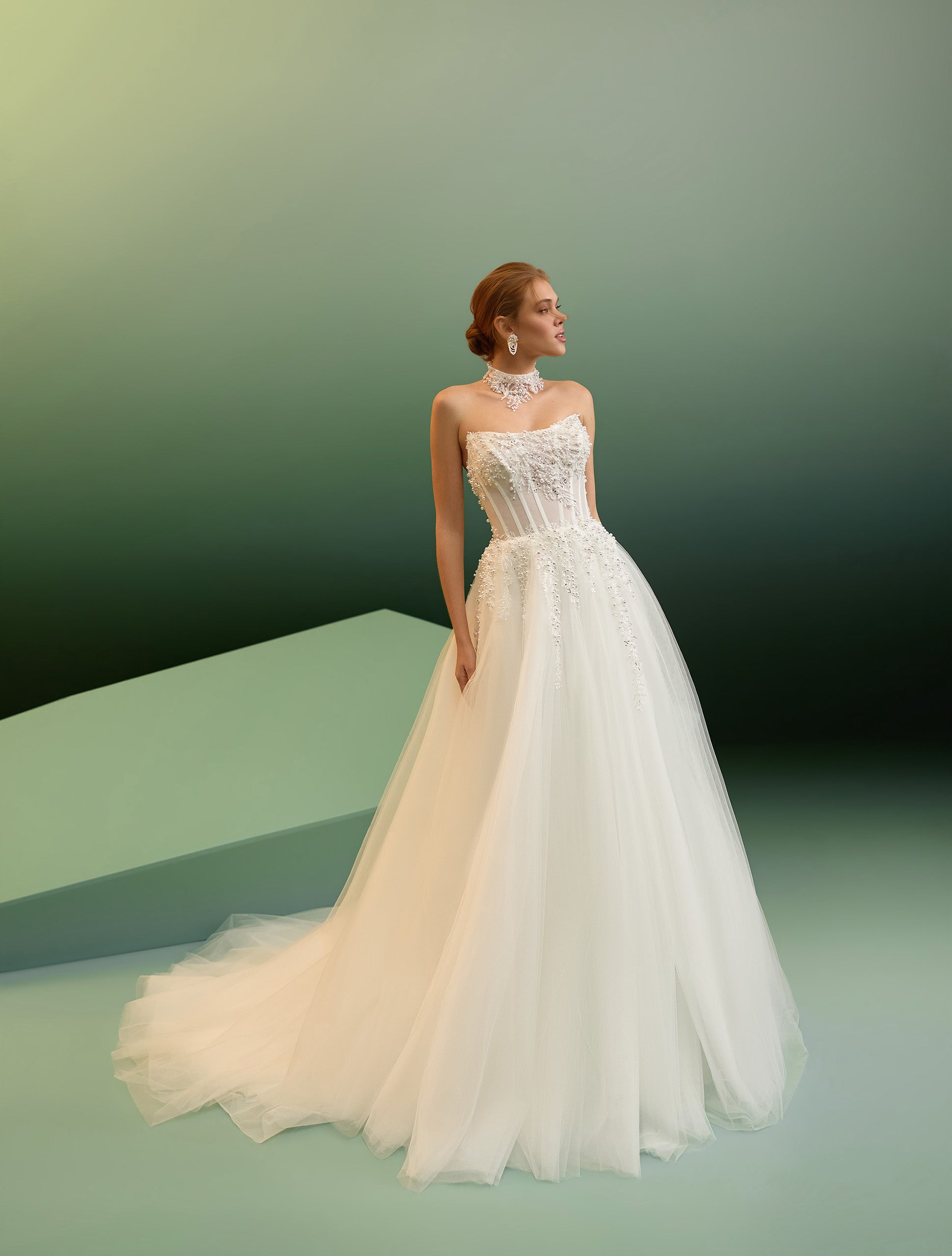 A woman modeling a strapless white wedding gown with a fitted embellished bodice and a full tulle skirt, posed against a soft green gradient studio background.
