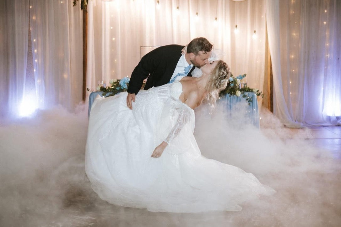 Couple in wedding attire embracing in a cloud of smoke with decorative lights and flowers in the background.